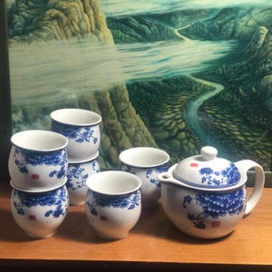 May include: A white porcelain tea set with blue floral designs. The set includes a teapot with a lid and six matching teacups. Each cup has a blue floral pattern and a small red square. The set is on a wooden surface.