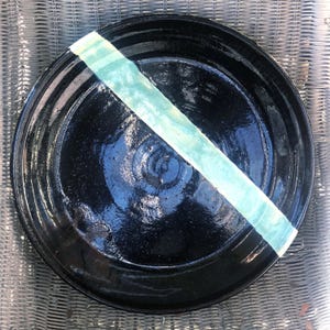 May include: A handmade black ceramic plate with a diagonal stripe of light blue and white glaze. The plate has a glossy finish and a slightly textured surface. This decorative plate is suitable for serving food.