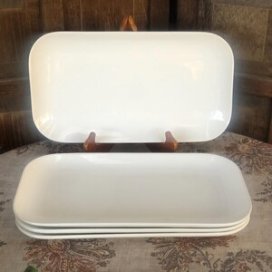 May include: A set of four rectangular white ceramic serving platters with rounded corners. The platters feature a smooth, glossy finish and a shallow depth, stacked neatly on top of each other, with one additional platter displayed upright on a stand.
