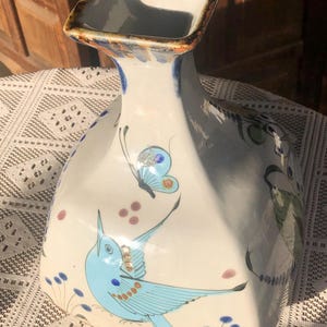 May include: A white ceramic vase with a square opening and a flared body. The vase is decorated with a blue bird, a butterfly, and floral accents. The rim has brown and orange accents.