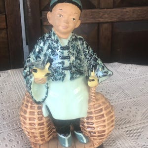 May include: A ceramic figurine of a boy wearing a blue hat and a green and blue patterned robe. He is holding two yellow birds in his hands and has two wicker baskets on his back.