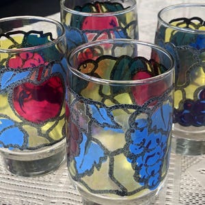 May include: Four clear drinking glasses with colorful fruit designs. Each glass features a different fruit design, including apples and grapes, with blue, red, and yellow accents. The designs are outlined in black.
