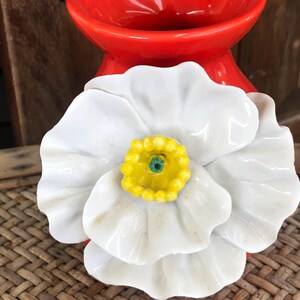May include: A white ceramic flower with a yellow and green center, set against a red ceramic vase. The flower has large, delicate petals. The background is a woven surface.