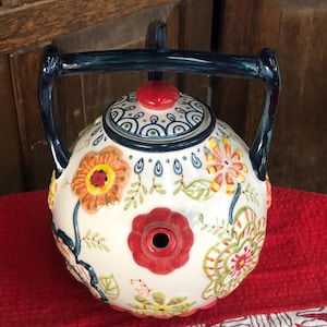May include: A white ceramic teapot with a floral design in orange, yellow, and green. The teapot has a red flower-shaped spout and a blue handle. The lid is topped with a red knob and decorated with blue and white patterns. The teapot is on a red surface.