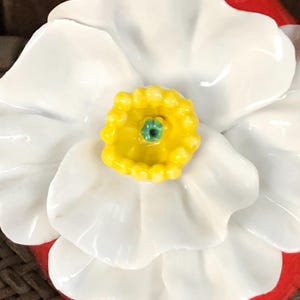 May include: Close-up of a ceramic flower with white petals and a yellow center with a green accent. The flower is set against a red base. The design is likely for home decor or a decorative item.