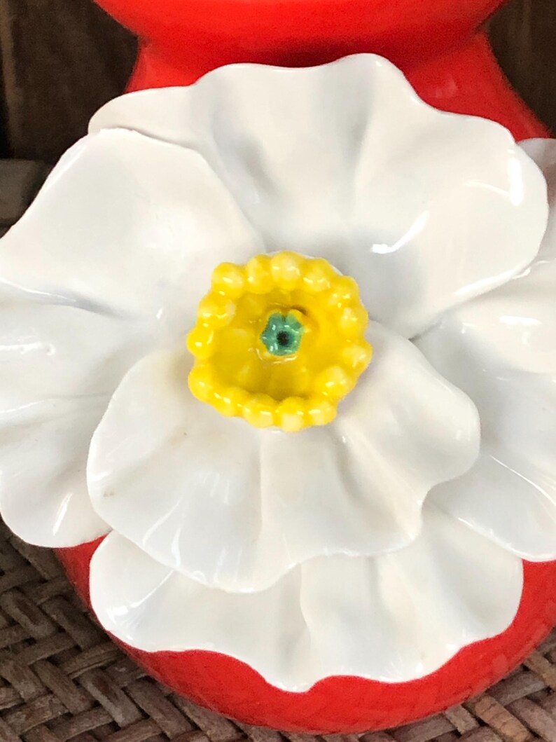 May include: Close-up of a ceramic flower with white petals and a yellow center with a green accent. The flower is set against a red base. The design is likely for home decor or a decorative item.