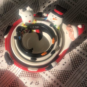 May include: A set of Christmas-themed measuring cups. The set includes a Santa Claus, a snowman, and a reindeer. The cups are white with red and black accents. The smallest cup is labeled "1/4 cup".