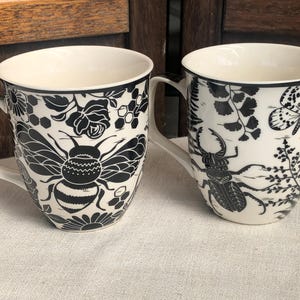 May include: Two white ceramic mugs with black insect and floral designs. One mug features a bee, the other a beetle, with floral and botanical accents. Both mugs have a black rim and white handle.