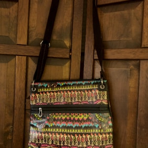 May include: A crossbody bag with a colorful, patterned design. The bag features a black background with a vibrant mix of pink, yellow, teal, and white geometric shapes. It has a black adjustable strap and a zippered front pocket.
