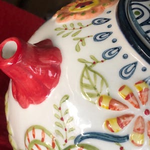 May include: Close-up of a white ceramic teapot with a red spout and handle. The teapot is decorated with colorful floral and leaf patterns in orange, yellow, green, and blue. The design has a raised, textured appearance.