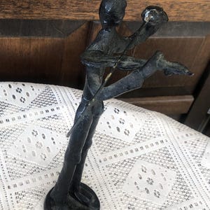 May include: A dark bronze sculpture of a person playing a stringed instrument. The figure is posed with one leg extended and arms raised, holding the instrument. The sculpture has a circular base and is set against a patterned white lace tablecloth.