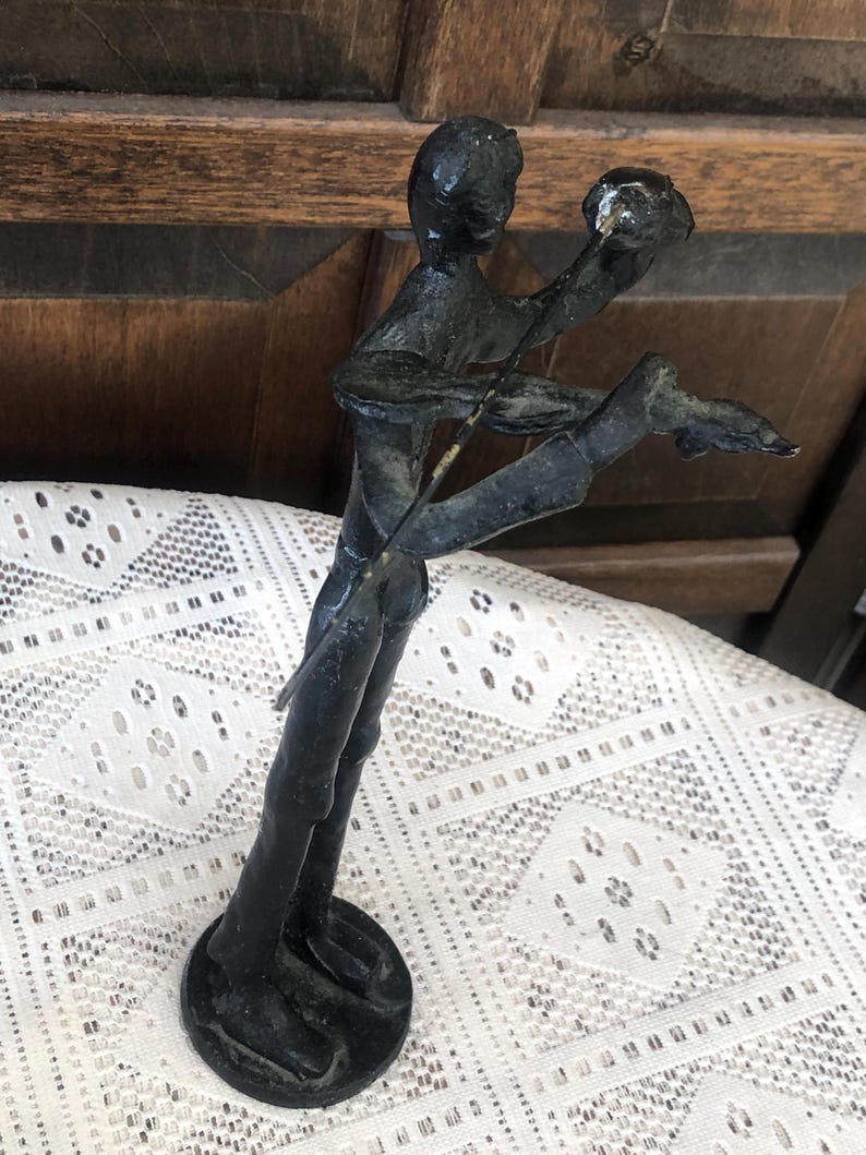 May include: A dark bronze sculpture of a person playing a stringed instrument. The figure is posed with one leg extended and arms raised, holding the instrument. The sculpture has a circular base and is set against a patterned white lace tablecloth.