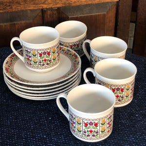 May include: A set of white ceramic teacups and saucers. The cups and saucers have a floral pattern with red, yellow, purple, and green flowers. The set includes six saucers and four cups.