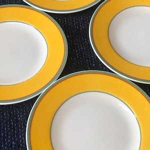 May include: Four white ceramic plates with a vibrant yellow border, accented by thin green and red lines. The plates are arranged on a dark blue textured surface, showcasing their bright design. The plates are a classic tableware item.