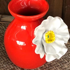 May include: A vibrant red ceramic vase with a glossy finish. A large white flower with a yellow center is attached to the side. The vase has a rounded body and a wide opening, suitable for floral arrangements.