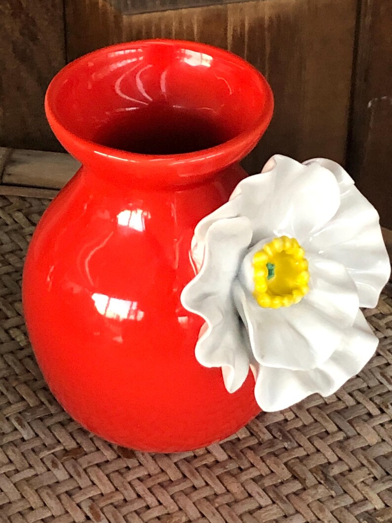 May include: A vibrant red ceramic vase with a glossy finish. A large white flower with a yellow center is attached to the side. The vase has a rounded body and a wide opening, suitable for floral arrangements.