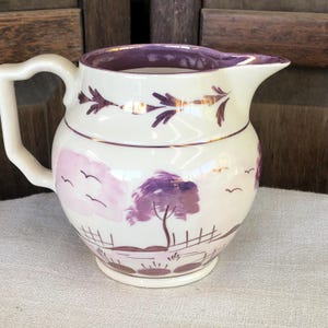 May include: Cream-colored ceramic pitcher with a purple landscape design. The pitcher features a purple rim and handle, with a gold accent band. The landscape includes a tree, fence, and birds, creating a vintage aesthetic.