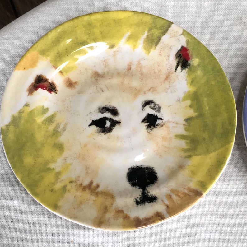 May include: A hand-painted ceramic plate with a dog's face design. The plate has a cream-coloured face with black features and a yellow-green border. A unique, artistic piece for the home.