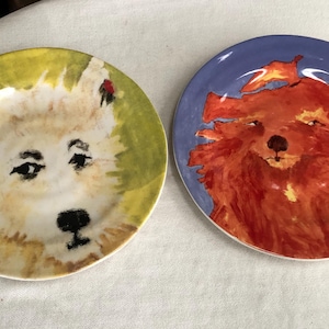 May include: Two decorative ceramic plates with dog portraits. One plate features a white dog with a black nose and eyes on a yellow and green background. The other plate has a red dog on a blue background.