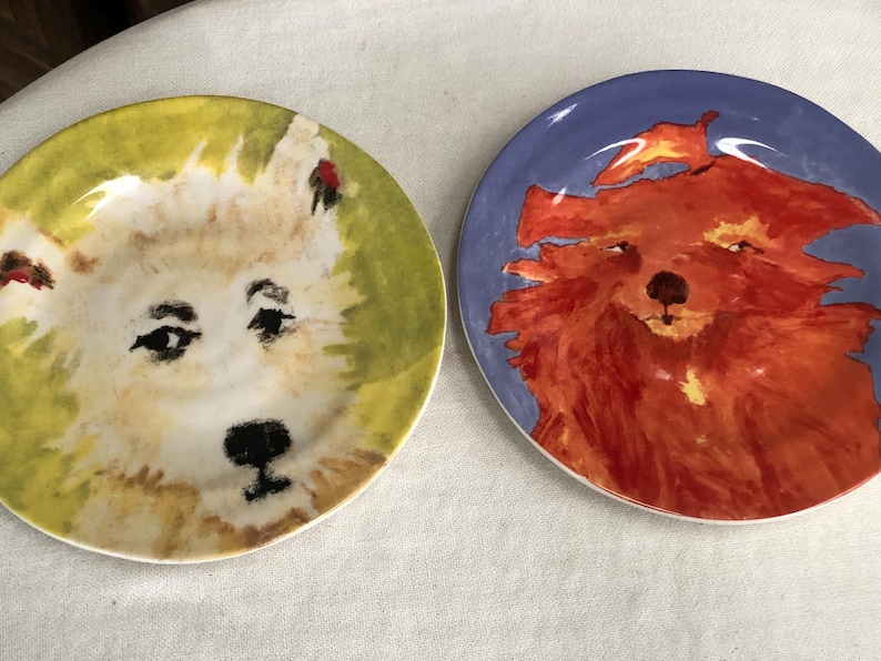 May include: Two decorative ceramic plates with dog portraits. One plate features a white dog with a black nose and eyes on a yellow and green background. The other plate has a red dog on a blue background.