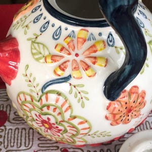 May include: A white ceramic teapot with a dark blue handle and rim. The teapot is decorated with colorful floral designs in red, orange, yellow, and green. The flowers are stylized and painted in a folk art style.