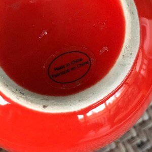 May include: A close-up of a bright red ceramic vase with a white rim. The base of the vase has a sticker that reads "Made in China / Fabriqu&eacute; en Chine". The vase has a glossy finish.