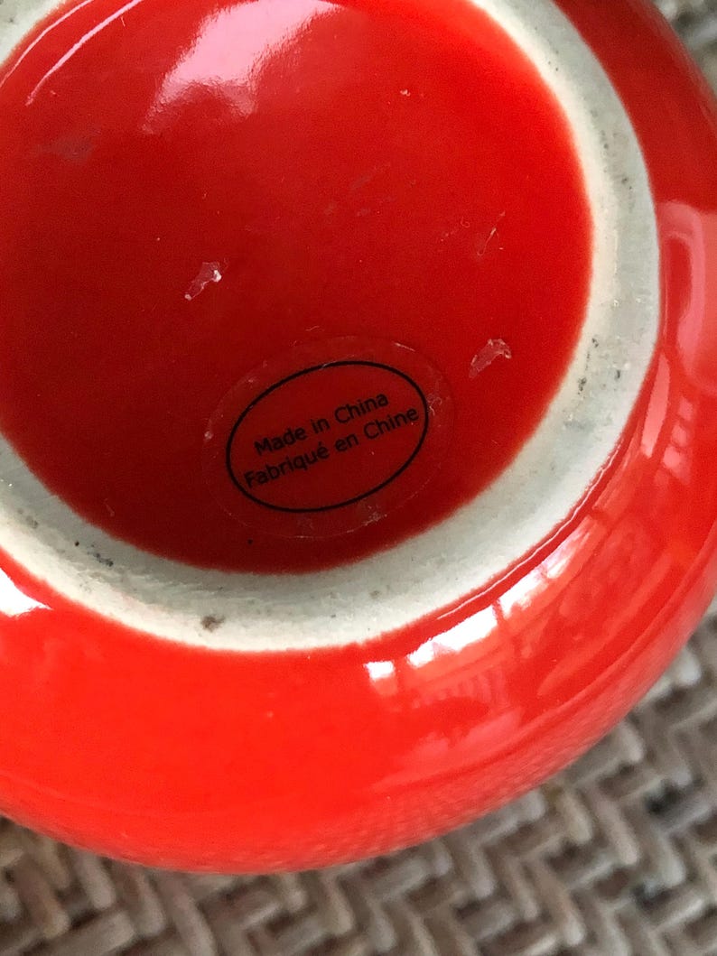 May include: A close-up of a bright red ceramic vase with a white rim. The base of the vase has a sticker that reads "Made in China / Fabriqu&eacute; en Chine". The vase has a glossy finish.
