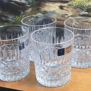 May include: Four clear, cut-crystal drinking glasses with a diamond and vertical line pattern. Each glass has a cylindrical shape and a thick base. The glasses are displayed on a wooden surface, with a painting in the background.