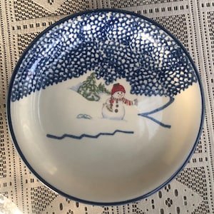 May include: A white ceramic plate with a blue rim and a winter scene. The plate features a snowman wearing a red hat and scarf, standing in the snow. Above the snowman is a blue dotted pattern, resembling a snowy sky.