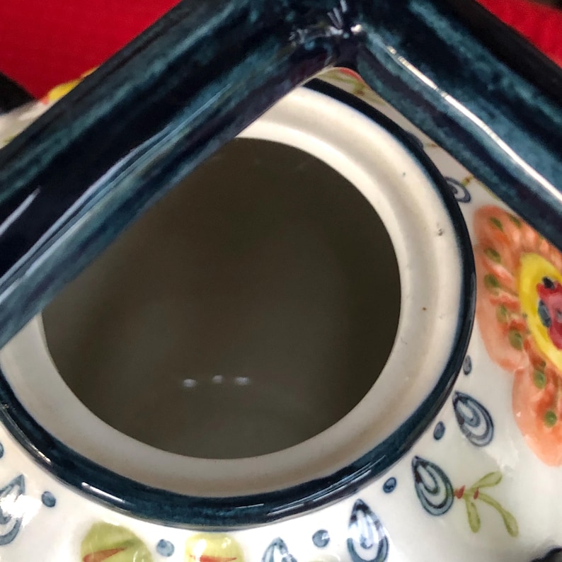 May include: A ceramic teapot with a dark blue handle and rim. The interior is a light gray color. The exterior is white with floral and leaf designs in orange, yellow, and blue. The teapot is set against a red background.
