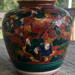 May include: A red and gold ceramic vase with a detailed design of people, trees, and fish. The vase has a wide mouth and a round body.