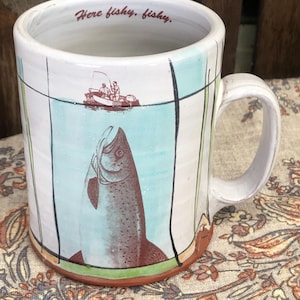 May include: A white ceramic mug with a handle, featuring a brown illustration of a fish being caught by a person in a boat. The mug has light blue and green accents with black vertical lines. The text Here fishy, fishy is printed on the inner rim.