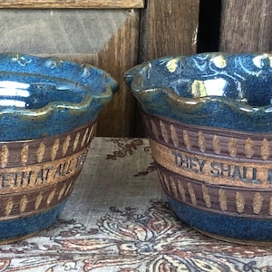May include: Two ceramic bowls with a glossy blue glaze on the interior and scalloped rims. The exterior features a natural brown clay band with vertical etched patterns and embossed text reading A FRIEND LOVETH AT ALL TIMES and THEY SHALL MOUNT UP WITH WINGS.