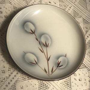 May include: A white ceramic dinner plate with a brown rim and a stylized design of a plant with three white cotton balls.