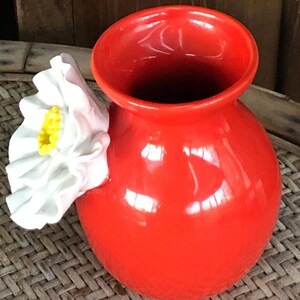 May include: A vibrant red ceramic vase with a glossy finish. A white flower with a yellow center is attached to the side. The vase has a rounded body and a wide opening. The vase is sitting on a woven surface.