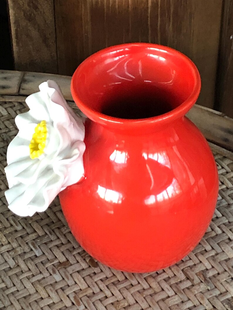 May include: A vibrant red ceramic vase with a glossy finish. A white flower with a yellow center is attached to the side. The vase has a rounded body and a wide opening. The vase is sitting on a woven surface.