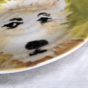 May include: A small, white ceramic plate with a hand-painted dog face design. The dog's face features black eyes, a black nose, and a light brown and yellow fur pattern. The plate's rim is white.