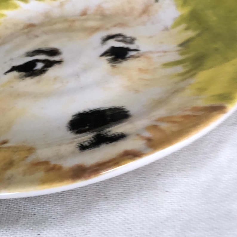 May include: A small, white ceramic plate with a hand-painted dog face design. The dog's face features black eyes, a black nose, and a light brown and yellow fur pattern. The plate's rim is white.