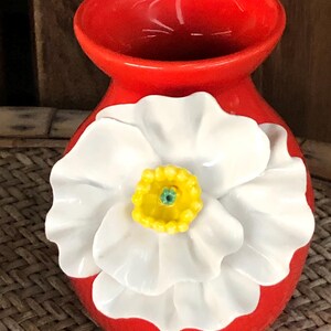 May include: A vibrant red ceramic vase with a large, white flower design. The flower has a yellow center with a green accent. The vase is a decorative piece, likely for displaying flowers or as a standalone accent.