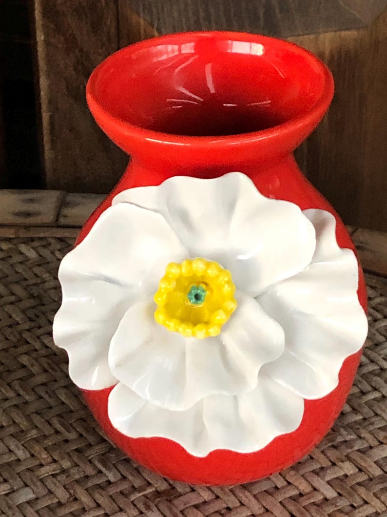 May include: A vibrant red ceramic vase with a large, white flower design. The flower has a yellow center with a green accent. The vase is a decorative piece, likely for displaying flowers or as a standalone accent.
