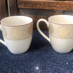 Puede incluir: Dos tazas de cerámica blanca con una banda color crema con círculos beige y un borde marrón fino. Cada taza tiene un asa curva y un interior blanco. Las tazas están sobre una superficie texturizada azul oscuro.