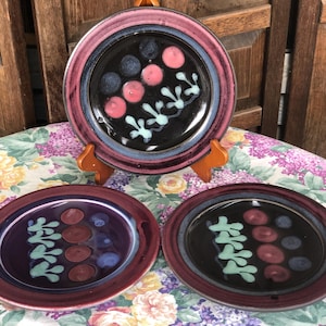 May include: Three decorative ceramic plates with a dark color scheme. The plates feature a black center with pink, blue, and white abstract designs. The rims are a mix of burgundy, pink, and blue. The plates are displayed on a floral tablecloth.