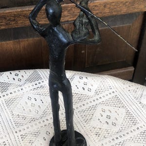 May include: A black metal sculpture of a person playing a violin. The figure is stylized with a simple form, holding the violin and bow. The sculpture is mounted on a round base and has a distressed finish.