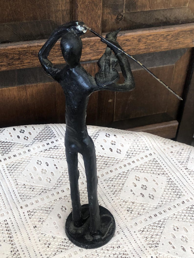May include: A black metal sculpture of a person playing a violin. The figure is stylized with a simple form, holding the violin and bow. The sculpture is mounted on a round base and has a distressed finish.