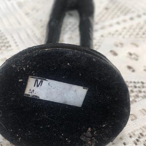 May include: A black sculpture of a figure with arms raised. The base is a dark, circular shape covered in a textured material. A white label with the letter "M" is affixed to the base.