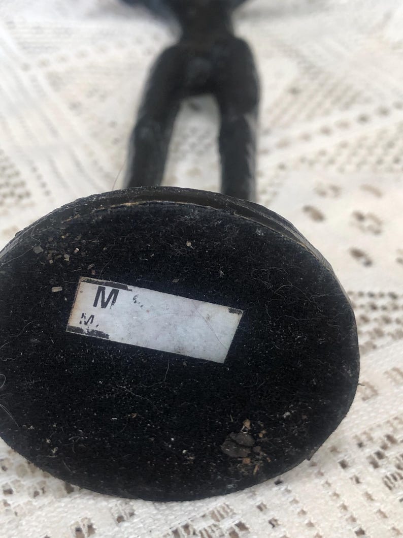 May include: A black sculpture of a figure with arms raised. The base is a dark, circular shape covered in a textured material. A white label with the letter "M" is affixed to the base.