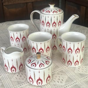May include: A white tea set with a red and gold geometric pattern. The set includes a teapot, creamer, sugar bowl, and four cups.