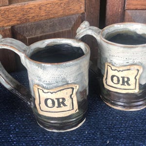 May include: Two handmade ceramic mugs with a gray and black glaze. Each mug has a handle and a rectangular label with the letters "OR" inside the outline of the state of Oregon.