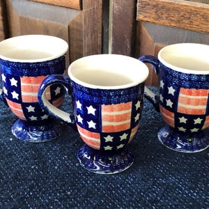 May include: Three ceramic mugs with a patriotic design. Each mug features a blue background with white stars and red and white stripes, resembling the American flag. The mugs have white interiors and handles, and sit on a pedestal base.