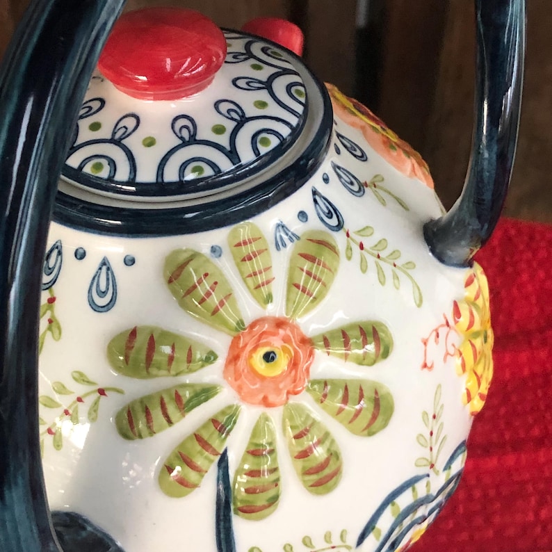 May include: A decorative ceramic teapot with a floral design. The teapot features a white base with a large flower in the center, green and red leaves, and a red lid. The handle and spout are dark blue.